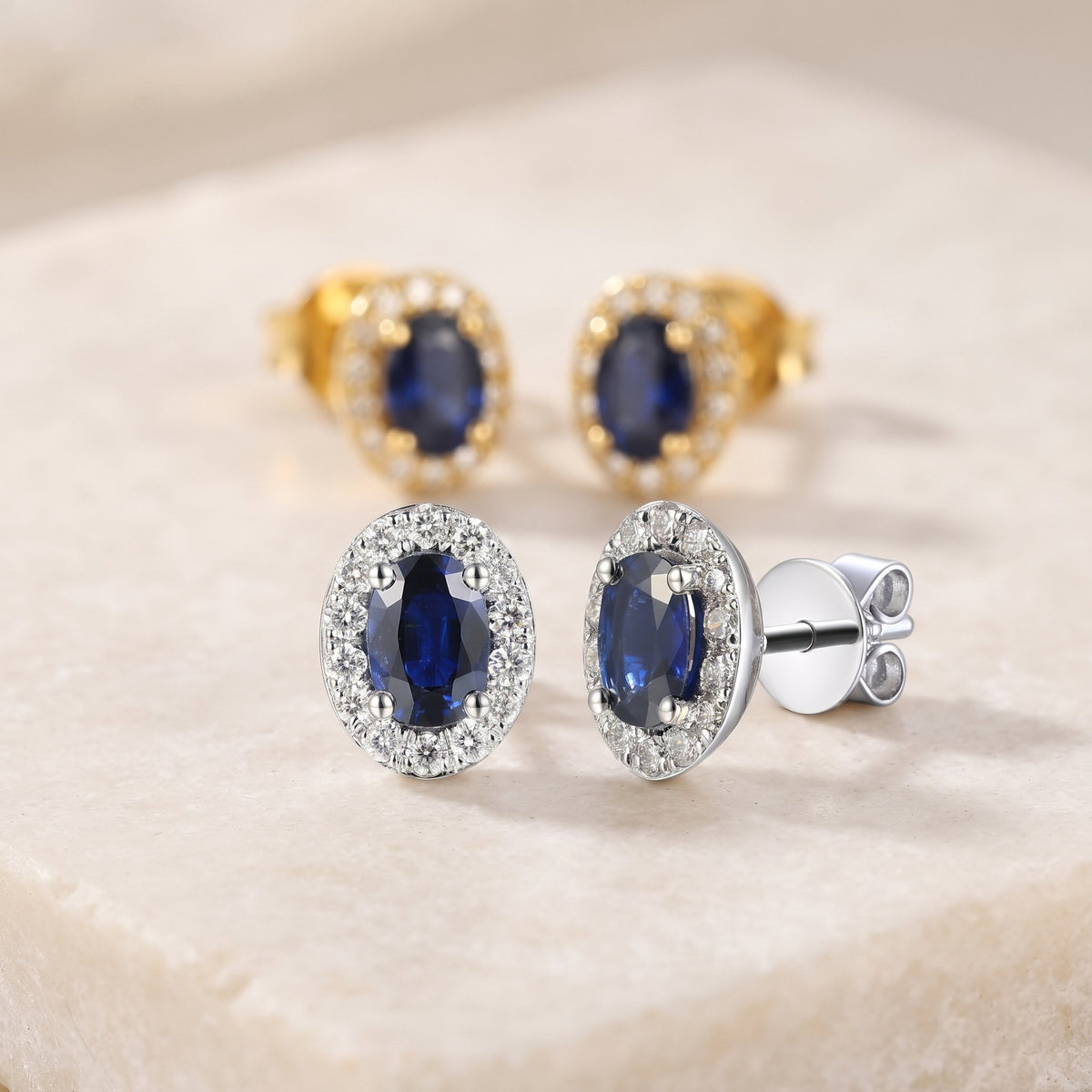 Oval blue sapphire and diamond halo earrings in 18k white gold and 18k yellow gold on a cream stone