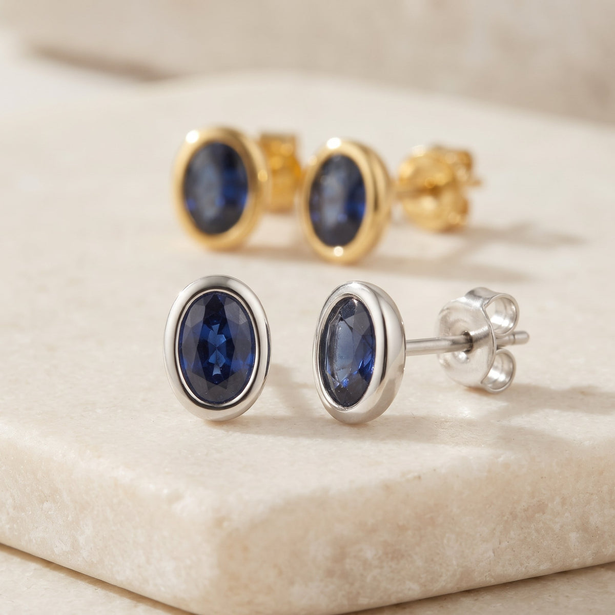 Oval blue sapphire rubover stud earrings in 18k white gold and 18k yellow gold on a cream stone