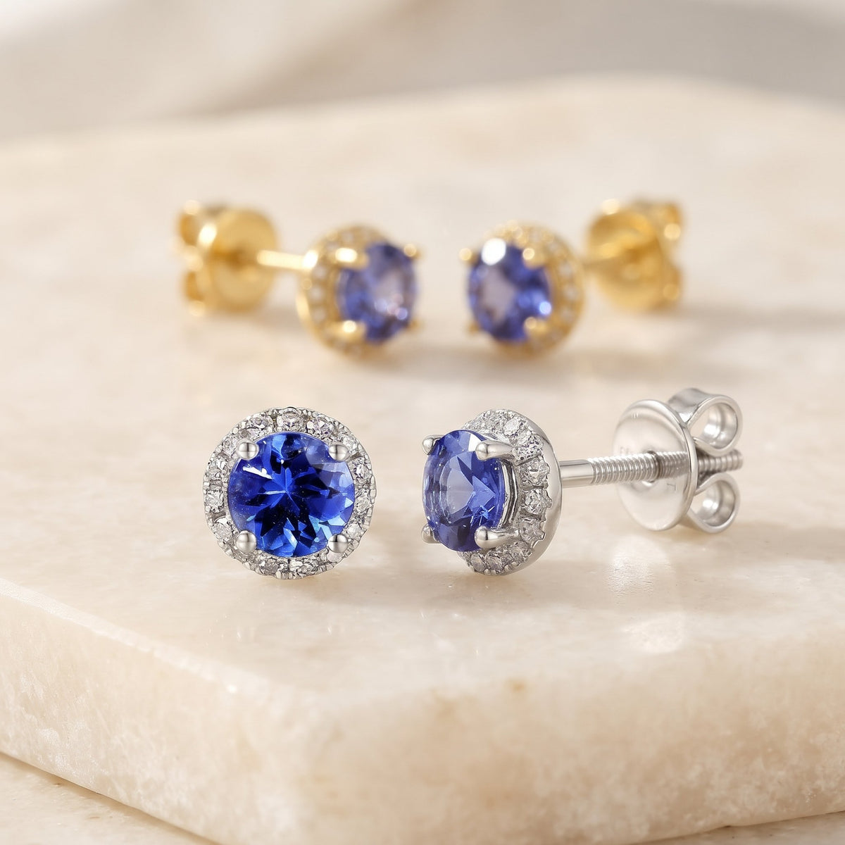 Round tanzanite and diamond halo earrings in 18k white gold and 18k yellow gold on a cream stone