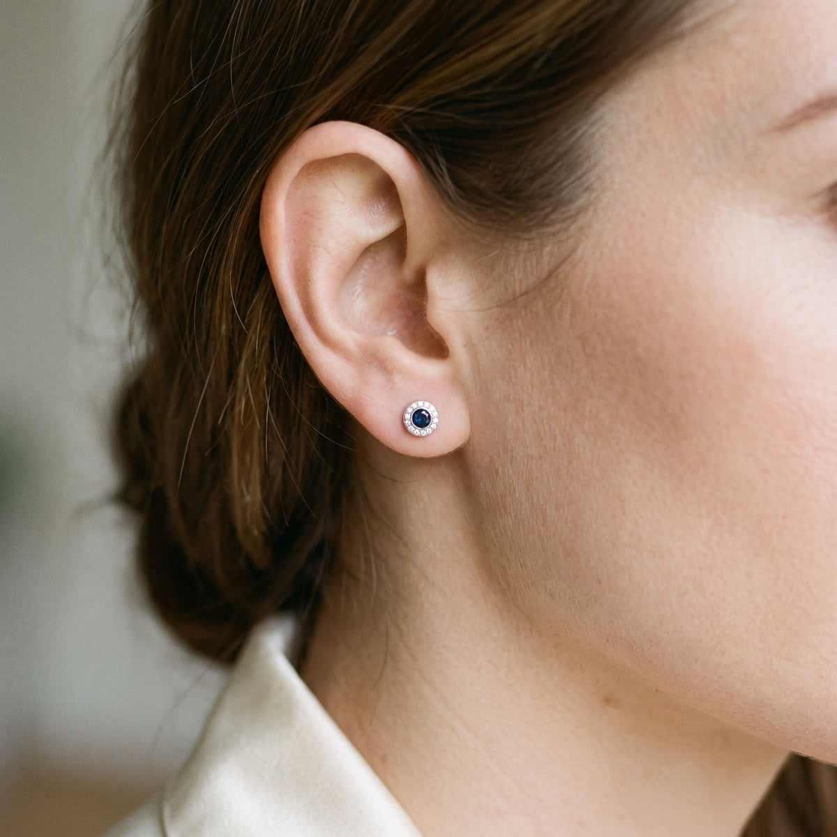 Round sapphire and diamond rubover halo stud earring worn on the ear, 18k white gold